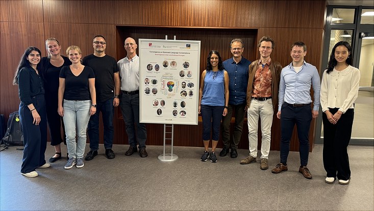 The CODILAC project leaders are standing in front of a wooden wall in a University building. At the centre, there's a poster stand showing the CODILAC project structure with pictures of the collaborating researchers.